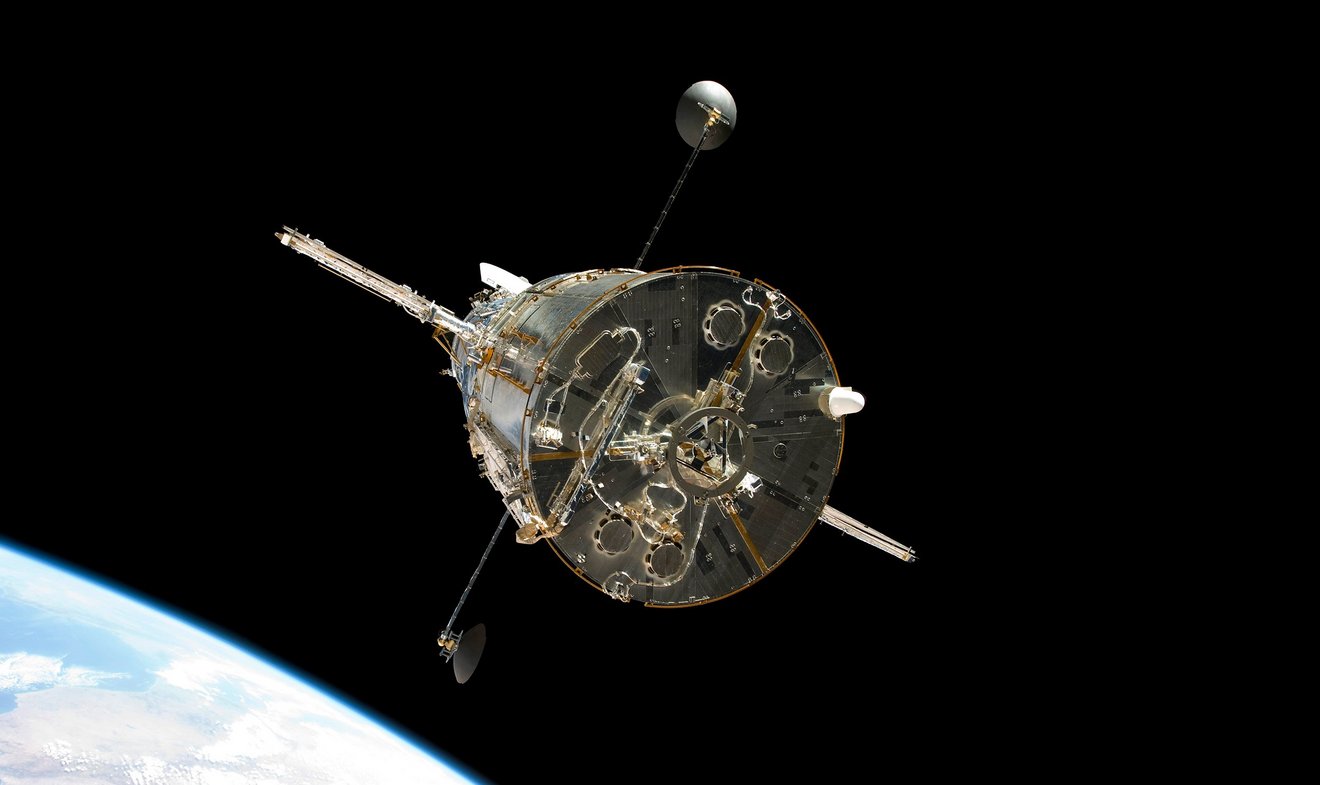 Picture of the telescope "Hubble" floating in space. A planet can be seen at the left edge of the image. The vehicle stands freely in the dark space. It is taken from below, antennas can be seen on the sides.