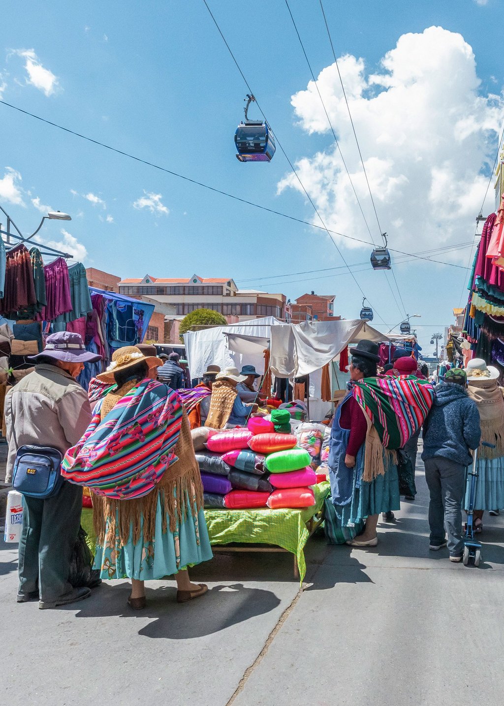 Photo of a market in La Paz, Bolivia. In the middle there is a stand with colorful fabrics where people stand. At the edges you can see stalls with hanging garments. The ropeway runs above the market situation.