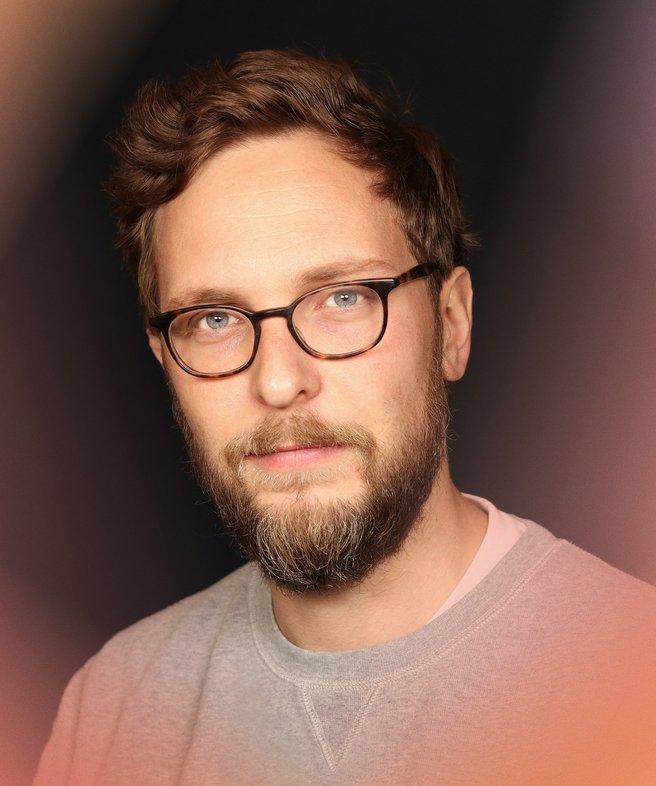 Portrait photo of Sascha Friesike with glasses and beard in a grey sweater looking into the camera from blue eyes.