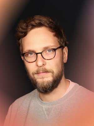 Portrait photo of Sascha Friesike with glasses and beard in a grey sweater looking into the camera from blue eyes.