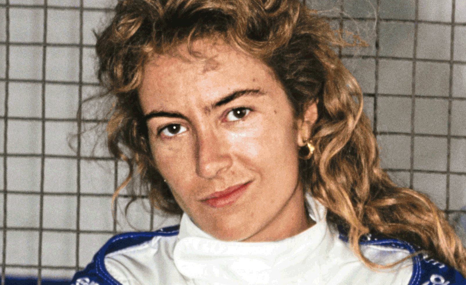 Portrait der Rennfahrerin Giovanna Amati, die in Fahrbekleidung vor einem Gitter in die Kamera blickt und lächelt.