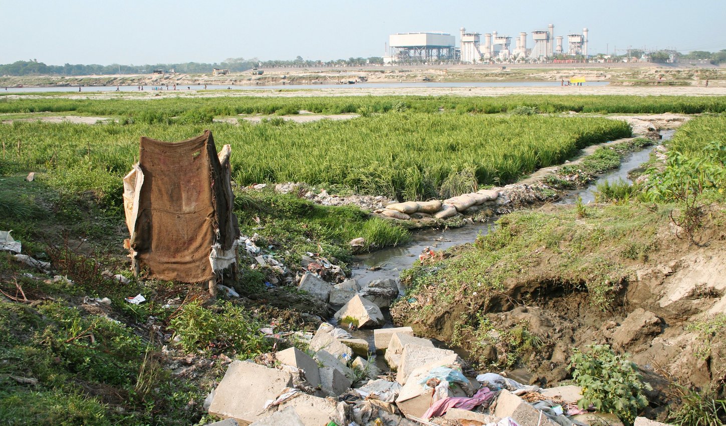 An einem kleine Bach auf einer grünen Wiese liegt viel Müll. Im Hintergrund sind an einem Fluss Menschen zu sehen.