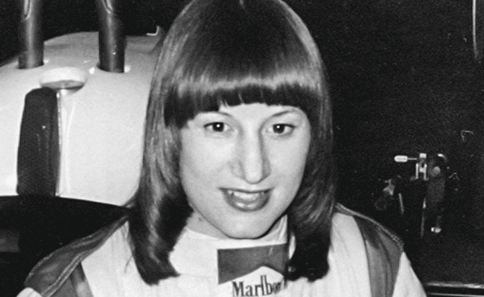 Portrait von Rennfahrerin Desiré Wilson, die in Fahrbekleidung in die Kamera lächelt.