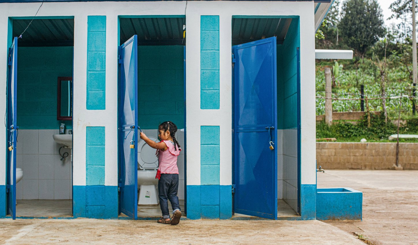 Ein kleines Mädchen schließt die Tür einer Toilettenanlage. Das balube Gebäude steht auf einem braunen Boden.