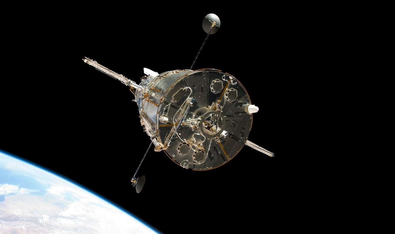 Aufnahme des Teleskopes "Hubble", das im Weltall schwebt. Am linken Bildrand ist ein Planet zu erkennen. Das Gefährt steht frei im dunkeln Raum. Es ist von unten aufgenommen, an den Seiten sind Antennen zu sehen.