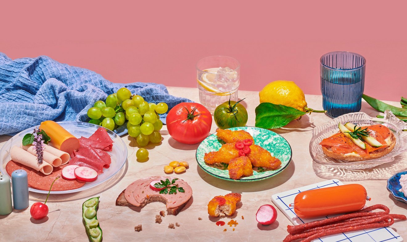 Foto von Lebensmitteln auf rosa Marmor vor altrosa Hintergrund. Natürliche und verarbeitete Lebensmittel nebeneinander. Obst, Gemüse und vegane Wurstwaren sind dekorativ angerichtet, daneben liegt ein blaues Küchentuch.