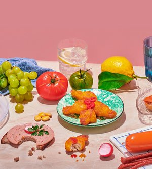 Foto von Lebensmitteln auf rosa Marmor vor altrosa Hintergrund. Natürliche und verarbeitete Lebensmittel nebeneinander. Obst, Gemüse und vegane Wurstwaren sind dekorativ angerichtet, daneben liegt ein blaues Küchentuch.
