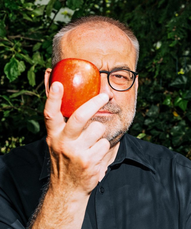 Portrait von Thomas Bauer, mit Brille, in dunkelblauem Hemd, der einen Apfel vor das rechte Auge hält.