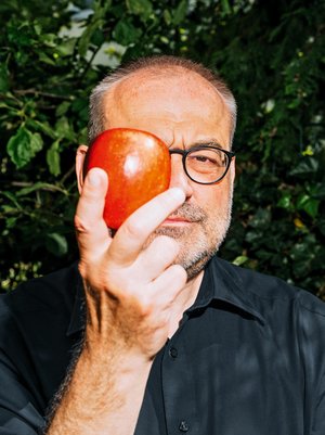 Portrait von Thomas Bauer, mit Brille, in dunkelblauem Hemd, der einen Apfel vor das rechte Auge hält.