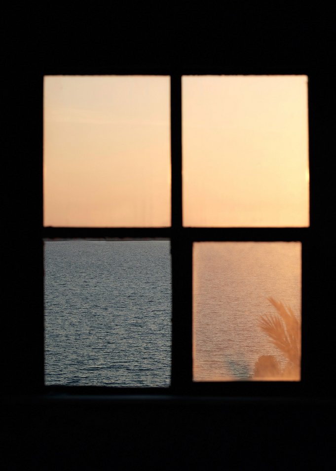 Foto eines Fensters aus dem Inneren eines Raumes aufgenommen. Drei der Fenstergläser sind gelblich-milchig und eines fehlt. Dort wo das Glas fehlt, wird der Blick auf ein Meer und seine wellige Wasseroberfläche frei.
