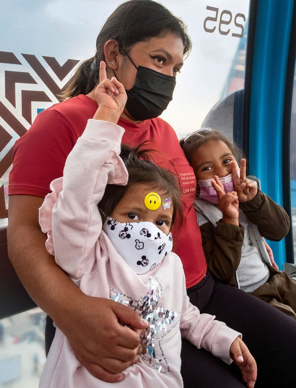 Close-up of three people in a gondola over Mexico City. A person sits in the wagon wearing a mask and holding two children in her arms. Both wear masks, look into the camera and form victory signs with their fingers.