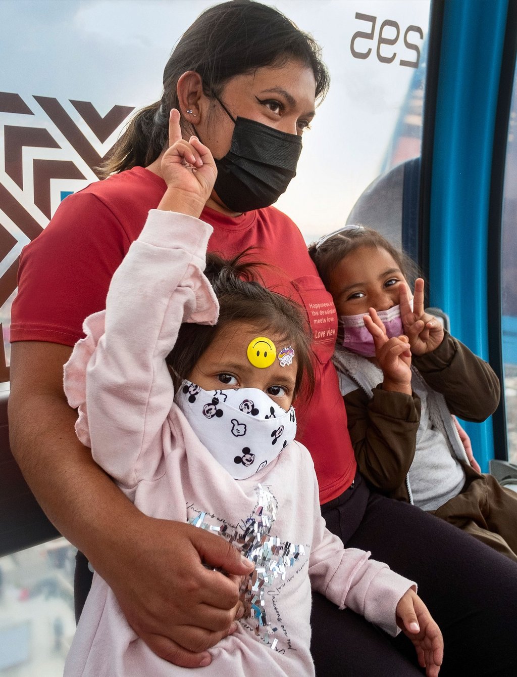 Nahaufnahme von drei Menschen in einer Gondel über Mexiko-Stadt. Eine Person sitzt in dem Wagon trägt eine Maske und hält zwei Kinder im Arm. Beide tragen Masken, blicken in die Kamera und formen mit ihren Fingern Victory-Zeichen.