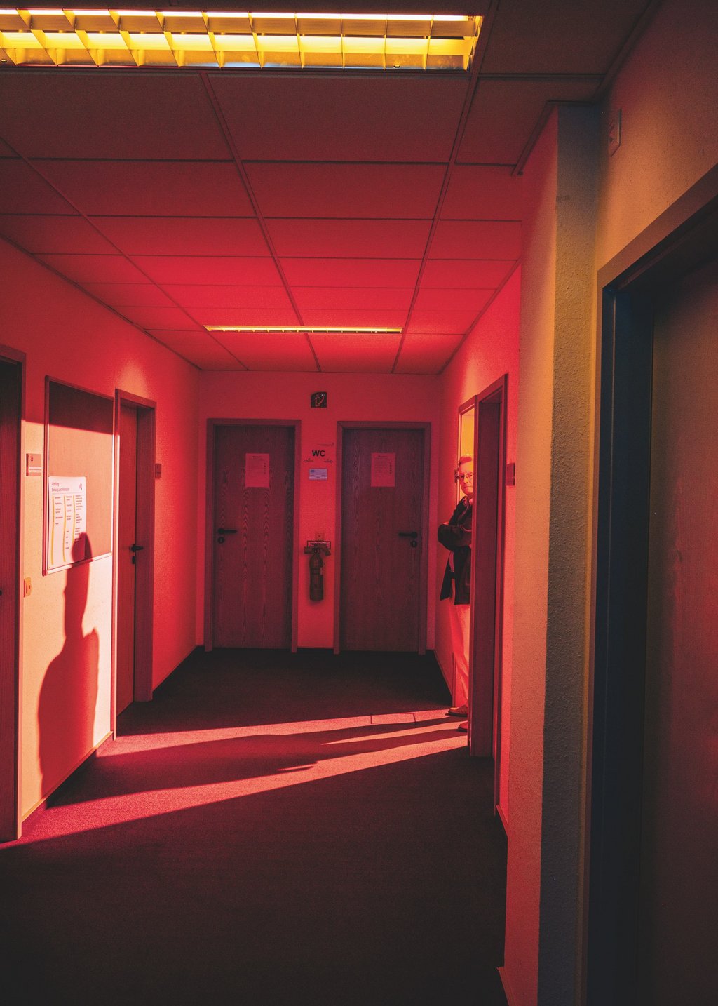 Ein rotbeleuchteter Flur mit vielen Türen. Eine Tür ist geöffnet, in ihr steht Michael Schetsche. Das Büro hinter der Tür leuchtet hell.