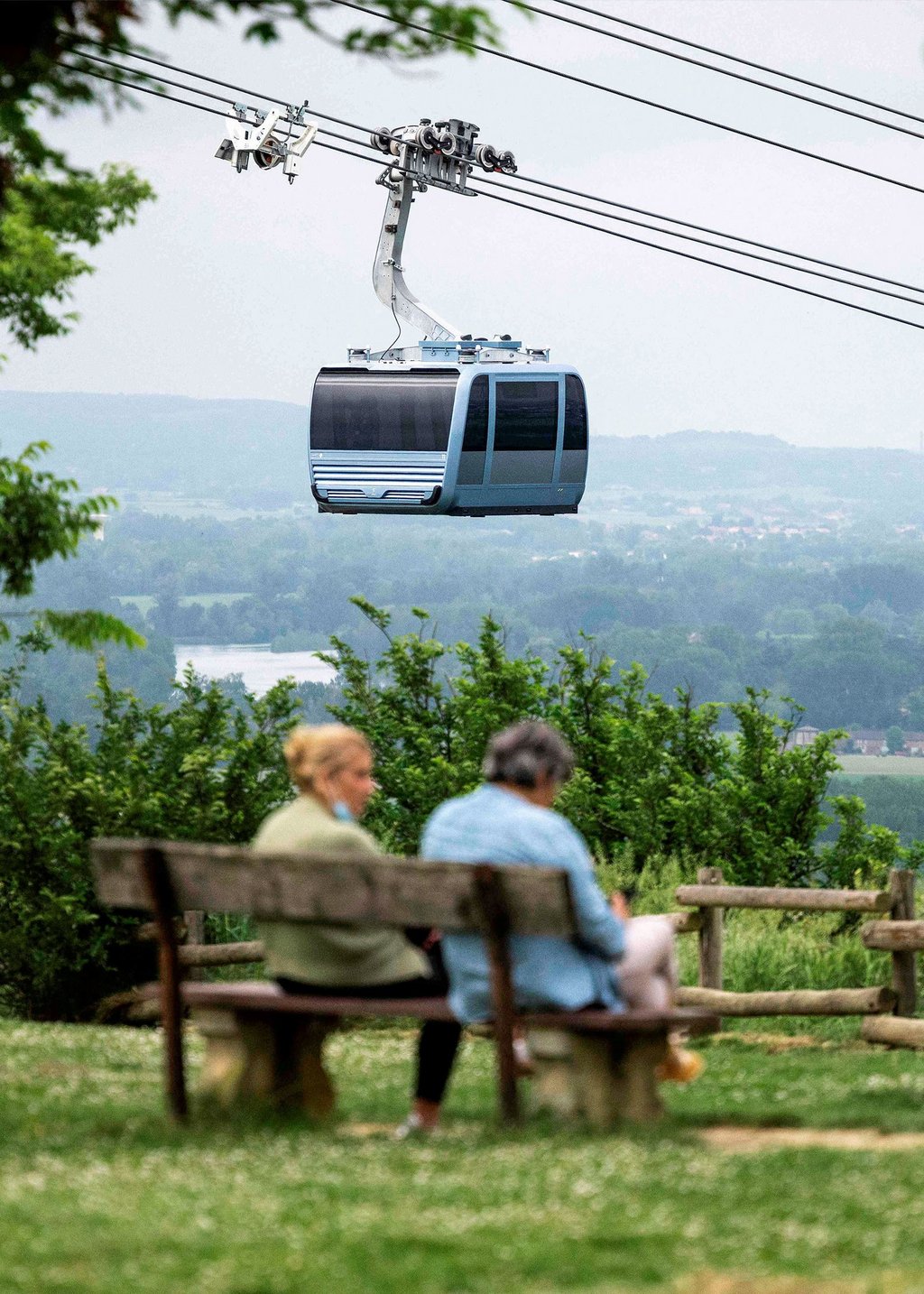 Photo of two people sitting on a bench on a mountainside in Toulouse, France, above which is a gondola of the Teleo ropeway.