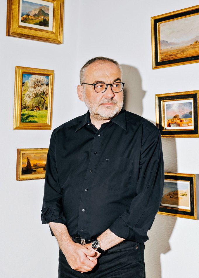 Thomas Bauer steht in einer Zimmerecke, an der Wand hängen viele gerahmte Landschaftsbilder. Er blickt in die Ferne.